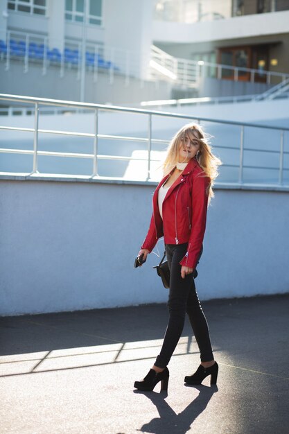 Stilvolle blonde Frau mit roter Jacke, die mit Sonnenlicht die Straße entlang geht