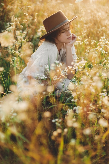 Stilvolle Boho-Frau mit Hut sitzt inmitten von Wildblumen im warmen Abendlicht. Sommerfreude und Reisen. Junge Frau in rustikalem Leinentuch entspannt sich auf der Sommerwiese. Stimmungsvoller Moment