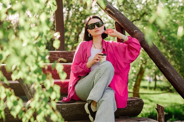 Stilvolle brünette Frau mit Sonnenbrille und Freizeitkleidung, die eine Tasse Kaffee hält und draußen im Park sitzt