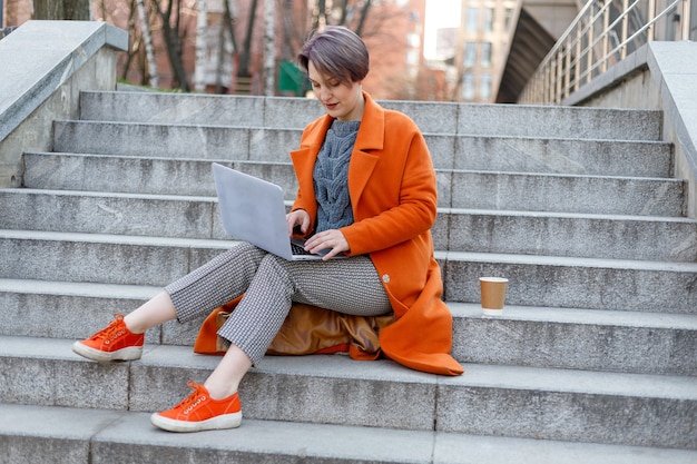 Stilvolle Dame im leuchtend orangefarbenen Mantel mit Laptop und Kaffeetasse