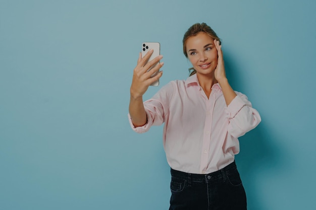 Stilvolle Dame in lässigem Hemd und Jeans macht Selfie-Pose, hält Smartphone und berührt sanft Gesicht isoliert auf blauem Hintergrund