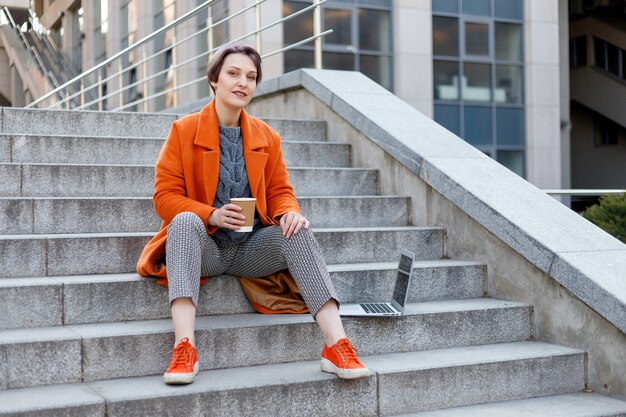 Stilvolle Dame in leuchtend orangefarbenem Mantel in einer Stadtszene, die einen Laptop benutzt und während einer Pause daran arbeitet. Outdoor-Technologie. Kaffee, Tee, Kakao trinken