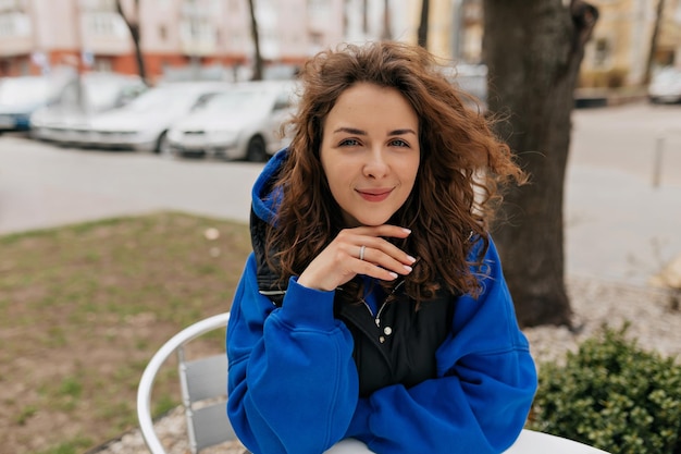 Stilvolle europäische Frau mit lockiger Frisur, die einen blauen Pullover trägt und in die Kamera posiert, während sie auf der Sommerterrasse in der Stadt sitzt Lifestyle-Porträt eines jungen kaukasischen Mädchens, das im Freien lacht