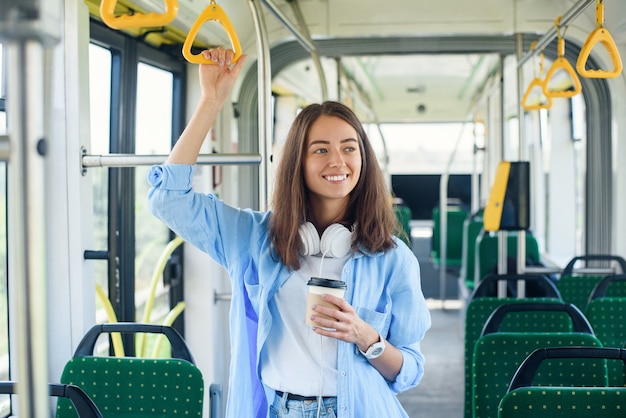 Stilvolle Frau im blauen Hemd, die Reise in der modernen Straßenbahn oder im Bus genießt, steht mit Tasse Kaffee im öffentlichen Verkehr.