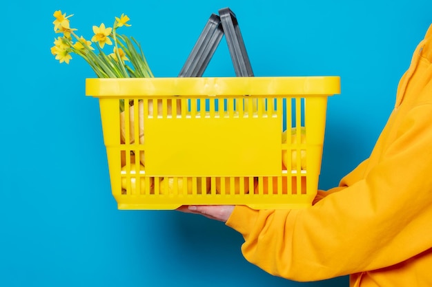 Stilvolle Frau im gelben Hoodie mit gelbem Korb und Blumen auf blauem Hintergrund