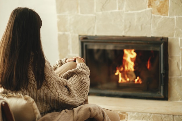 Stilvolle Frau im gemütlichen Pullover, die sich im Stuhl am Kamin aufwärmtHeizhaus mit Holzofen