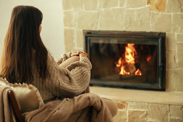 Stilvolle Frau im gemütlichen Pullover, die sich im Stuhl am Kamin aufwärmtHeizhaus mit Holzofen