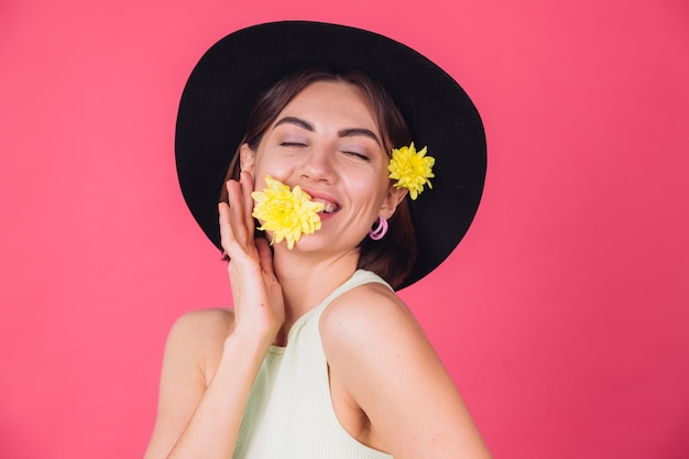 Stilvolle Frau im Hut, lächelnd mit zwei gelben Astern, niedlich halten eine Blume in Mundfrühlingsstimmung, glückliche Gefühle isolierten Raum