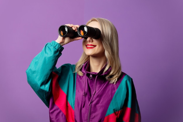 Stilvolle Frau in 80er Windjacke und roud Sonnenbrille hält Fernglas auf lila Wand