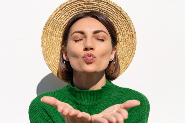 Stilvolle Frau in grünem lässigem Pullover und Hut im Freien auf weißer Wand ruhig und niedlich senden Luftkuss, der Sonnenstrahlen fängt