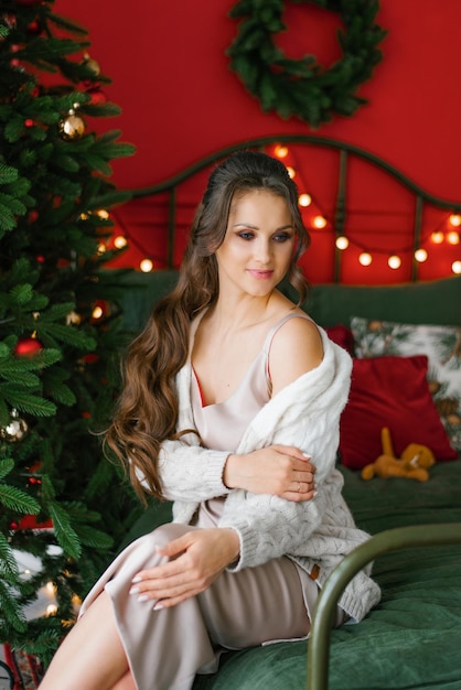 Stilvolle Frau in Hemd und Strickjacke sitzt auf dem Bett neben dem Weihnachtsbaum im Schlafzimmer