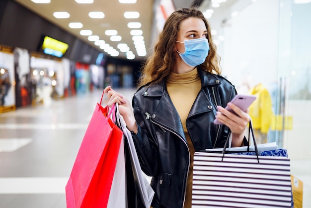 Stilvolle Frau in medizinischer Schutzmaske mit Einkaufstasche mit ihrem Telefon Modestil Blackfriday