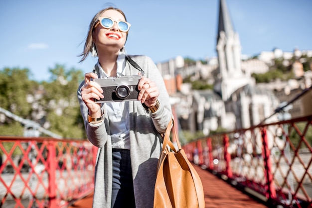 Stilvolle Frau mit Fotokamera, die die Morgenstadt genießt, die auf der berühmten roten Fußgängerbrücke in Lyon, Frankreich steht