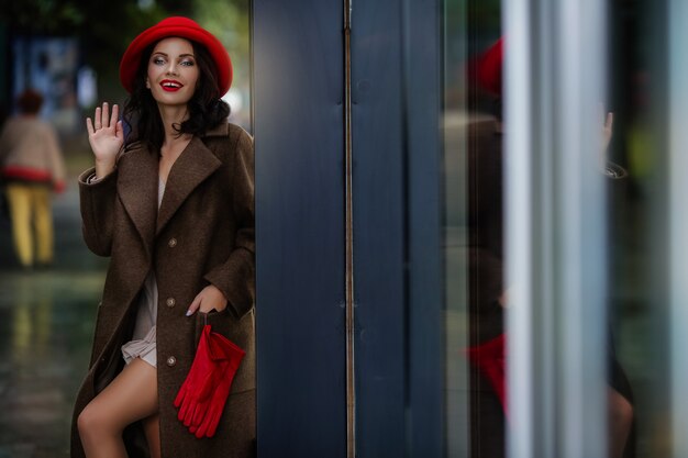 Stilvolle Frau mit hellem Abendmake-up in den roten Handschuhen und in einem roten Abendhut in der Stadt in einem Herbstmantel