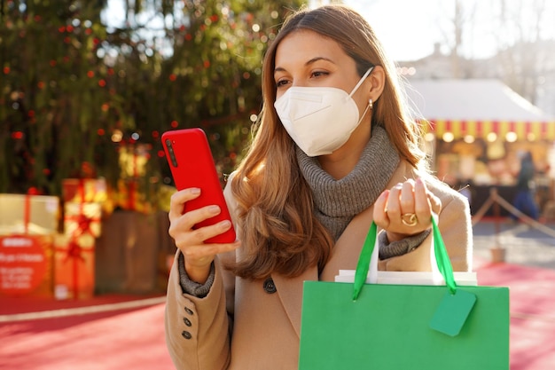 Stilvolle Frau mit KN95 FFP2-Gesichtsmaske, die ihr Telefon mit Einkaufstüten beobachtet, die zwischen den Weihnachtsmärkten spazieren gehen
