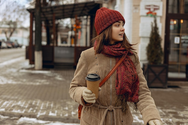 Stilvolle Frau mit warmem Hut und Mantel mit einer Tasse Kaffee zum Mitnehmen, die im Winter die Straße in der Stadt überquert