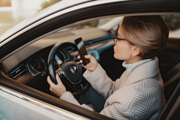 Stilvolle Frau sitzt im Auto gekleidet im Wintermantel und Brille mit Smartphone