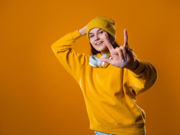 Stilvolle, fröhliche junge Brünette in einem gelben Hoodie und Hüten zeigt eine coole Geste mit zwei Fingern...