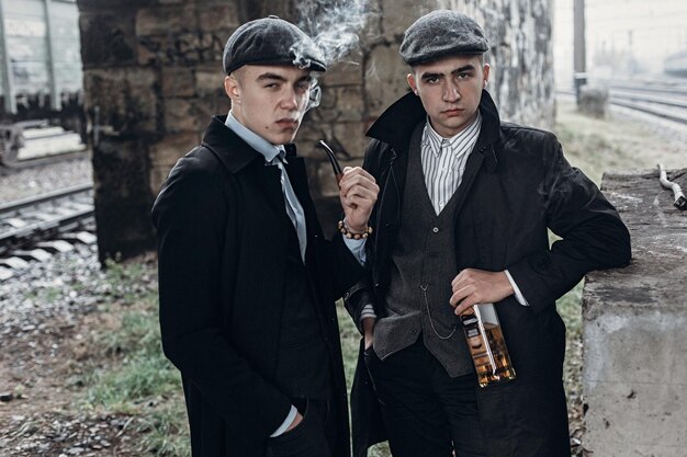 Stilvolle Gangstermänner, die auf dem Hintergrund der Eisenbahn rauchen, mit einer Flasche Alkohol, Sherlock Holmes und Doctor Watson England im Thema der 1920er Jahre
