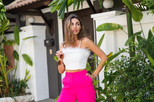 Stilvolle, gebräunte, schöne Frau mit Sonnenbrille, modischer rosa Hose und weißem Oberteil, die im Freien in einer luxuriösen tropischen Villa posiert