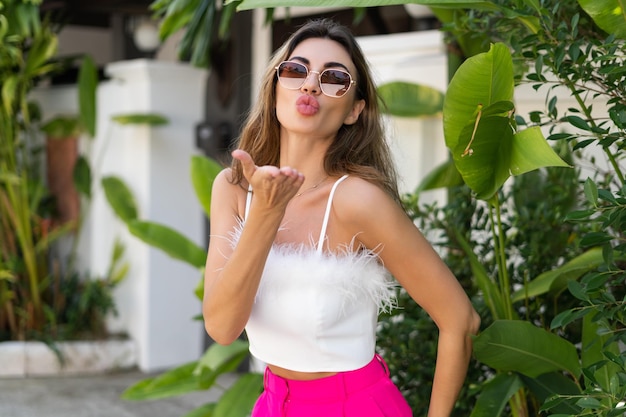 Stilvolle, gebräunte, schöne Frau mit Sonnenbrille, modischer rosa Hose und weißem Oberteil, die im Freien in einer luxuriösen tropischen Villa posiert