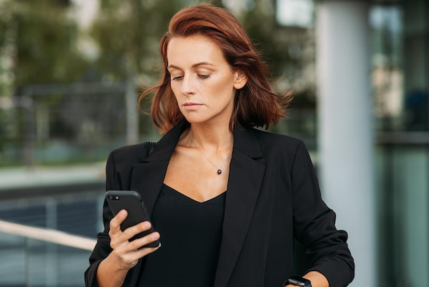 Stilvolle Geschäftsfrau mit roten Haaren, die schwarze formelle Kleidung trägt und auf das Mobiltelefon schaut