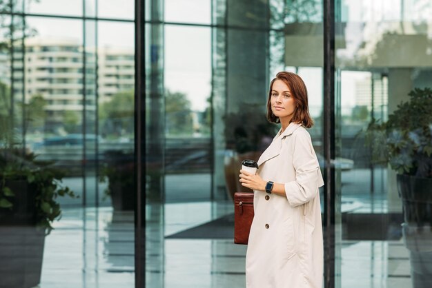 Stilvolle Geschäftsfrau steht bei einem Glasgebäude und hält einen Kaffee und einen Ordner