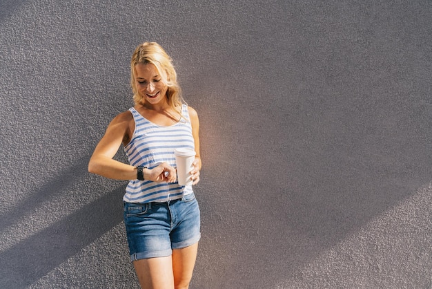 Stilvolle, glückliche junge Frau, die auf ihre Uhr schaut. Sie hält Papierkaffee zum Mitnehmen und genießt das Sonnenlicht in der Stadt. Porträt eines lächelnden Mädchens, Lifestyle-Konzeptbild.