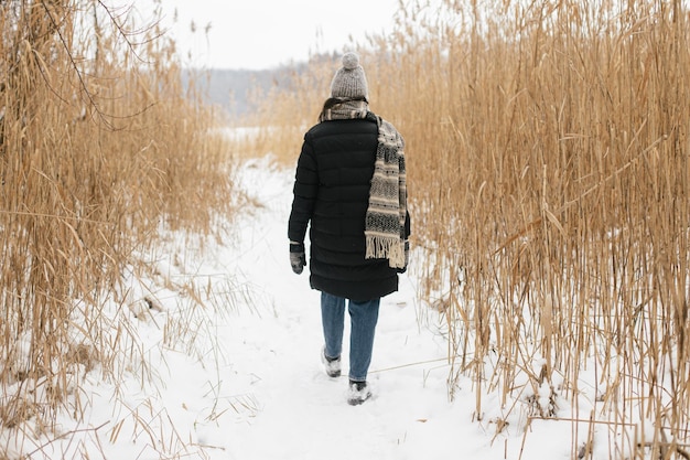 Stilvolle Hipster-Frau, die auf dem Hintergrund von getrocknetem Gras am verschneiten Wintersee Wanderlust geht