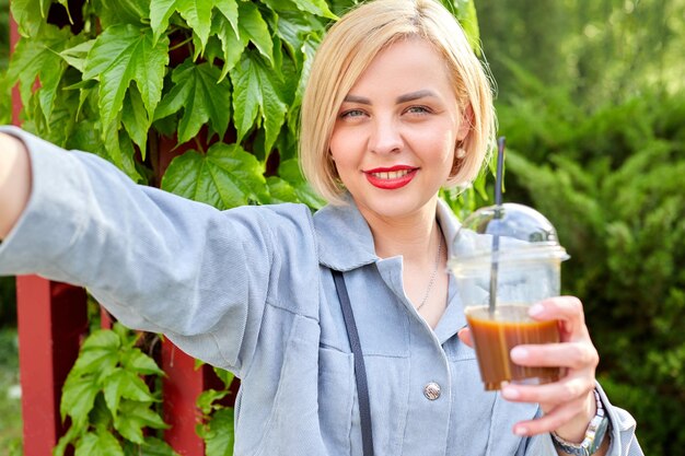 Foto stilvolle junge blondine läuft durch die stadt, trinkt ein kaltes getränk und macht ein selfie mit ihrem handy