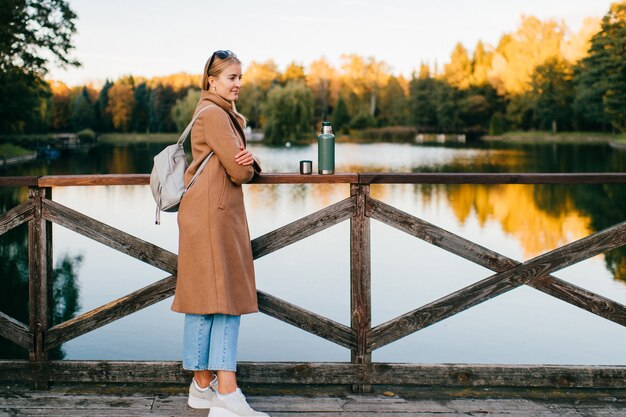 Stilvolle junge Dame im Mantel am Pier mit Thermoskanne