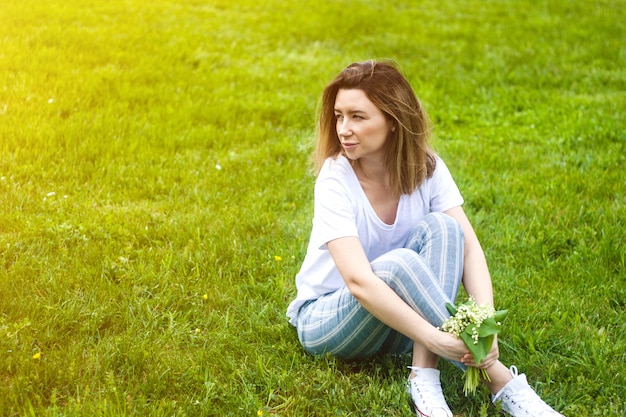 Stilvolle junge Frau, die auf grünem Gras am sonnigen warmen Tag sitzt