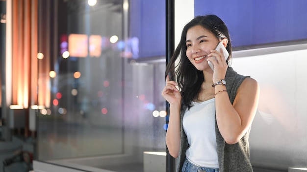 Stilvolle junge Frau, die mit dem Handy telefoniert, während sie auf der Bank vor der Vitrine des Einkaufszentrums sitzt