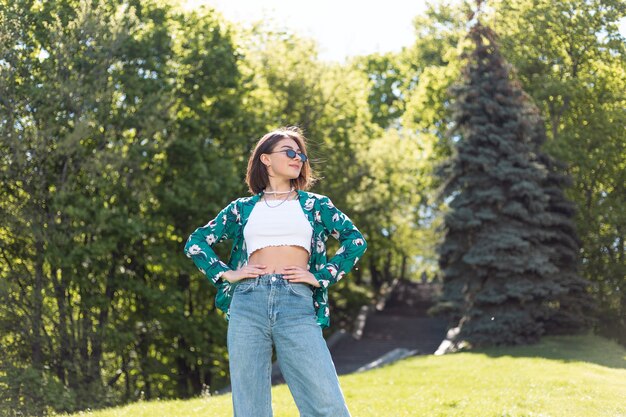 Stilvolle junge Frau im lässigen grünen Hemd und in den Jeans am sonnigen Tag, der auf Gras im Park aufwirft