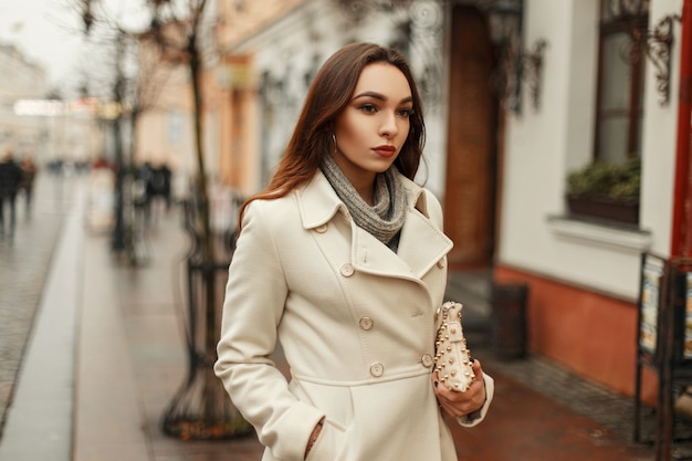 Stilvolle junge Frau im trendigen Herbstmantel mit Handtasche, die draußen in der Stadt geht