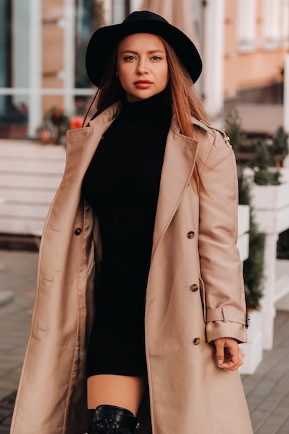 Stilvolle junge Frau in einem beigen Mantel in einem schwarzen Hut auf einer Stadtstraße