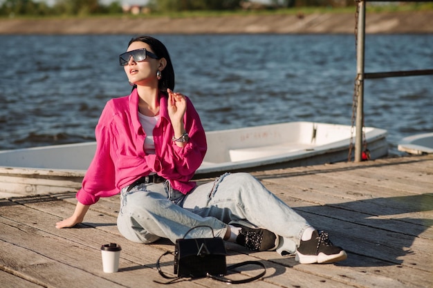 Stilvolle junge Frau in legerer Kleidung mit Sonnenbrille, die an einem sonnigen Tag auf einem Holzsteg sitzt