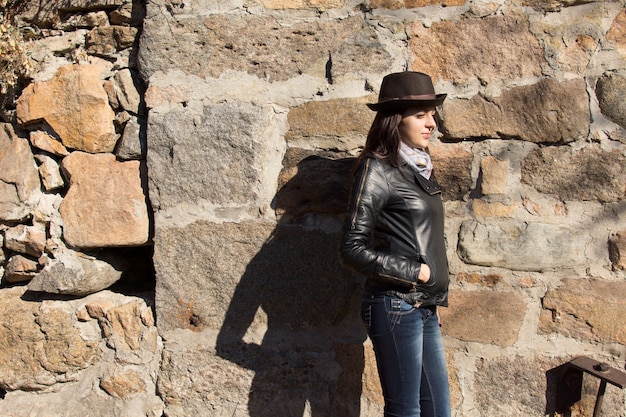 Stilvolle junge Frau in schwarzer Lederjacke, trendiger Mütze und Schal, die seitlich an einer alten Steinmauer im Sonnenschein mit Schatten steht