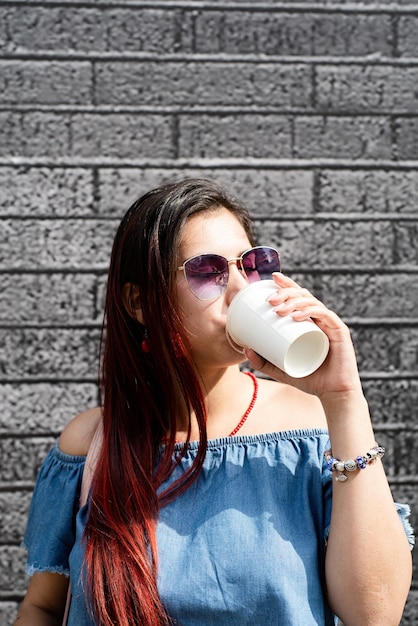 Stilvolle junge Frau mit Jeanshemd, Sonnenbrille und Tasche, die Kaffee zum Mitnehmen auf der Straße auf schwarzem Backsteinmauerhintergrund trinkt