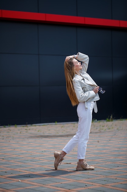 Stilvolle junge Frau mit langen blonden Haaren europäischen Aussehens mit einem Lächeln im Gesicht. Mädchen in weißer Jacke und weißer Jeans an einem warmen sonnigen Sommertag auf dem Hintergrund eines grauen Gebäudes