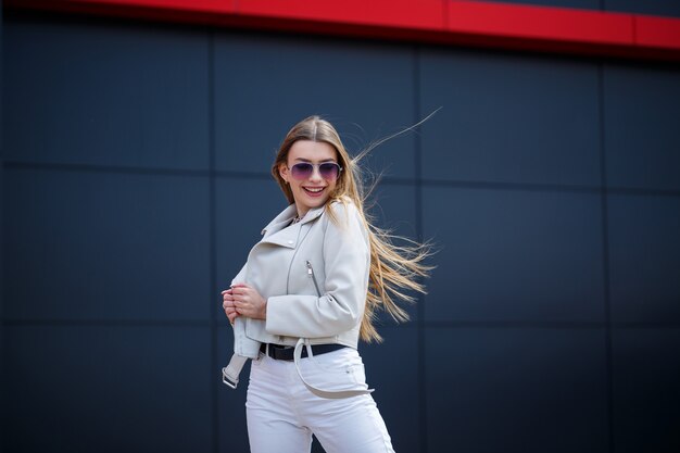 Stilvolle junge Frau mit langen blonden Haaren europäischen Aussehens mit einem Lächeln im Gesicht. Mädchen in weißer Jacke und weißer Jeans an einem warmen sonnigen Sommertag auf dem Hintergrund eines grauen Gebäudes