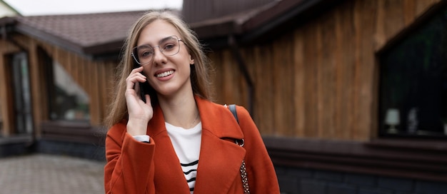 Stilvolle junge Geschäftsfrau spricht auf einem Mobiltelefon in einem leuchtend orangefarbenen Mantel auf dem Hintergrund von a