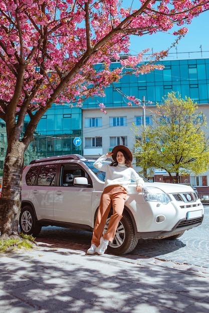 Stilvolle, lächelnde, schöne Frau, die neben dem blühenden Sakura-Baum des Autos auf dem Hintergrund steht, der Frühling kommt