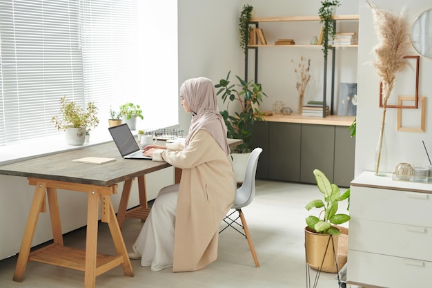 Stilvolle muslimische Frau mit Hijab sitzt alleine in einem gemütlichen Bürozimmer und arbeitet an ihrem Laptop-Kopierplatz