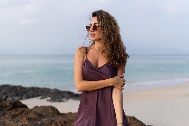Stilvolle, romantische, zarte, sinnliche Frau in Seidenkleid und Sonnenbrille am Strand bei Sonnenuntergang