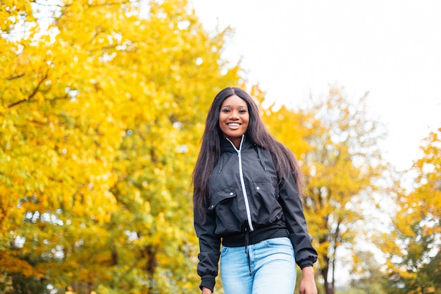 Stilvolle schöne junge Afroamerikanerin mit einem Lächeln in modischer Kleidung geht im Herbstpark mit leuchtend gelbem Herbstlaub spazieren