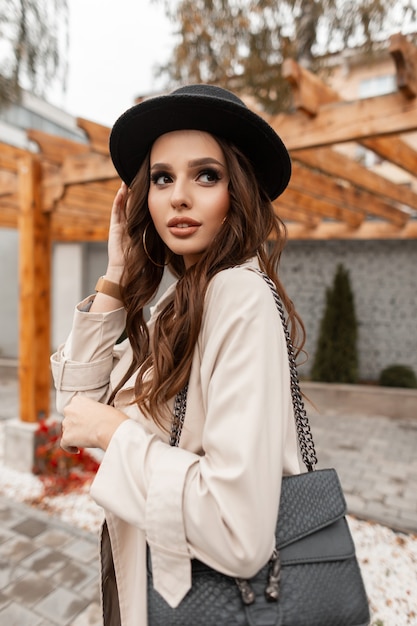 Stilvolle schöne junge Frau in modischer eleganter Kleidung mit Mantel, Hut und Lederhandtasche geht in der Stadt spazieren. Glamouröser femininer Stil und Schönheit