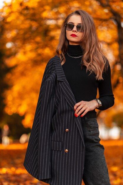 Stilvolle schöne junge Frau in modischer Kleidung mit Jacke, Pullover und Sonnenbrille posiert in einem Park mit hellem Herbstlaub. Weiblicher Geschäftsstil und Schönheit