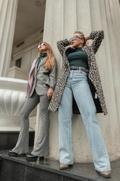 Stilvolle, schöne junge modische Frauen in modischer Kleidung mit Mänteln, Vintage-Jeansschal und Anzug, die in der Nähe von Retro-Säulen auf der Straße stehen
