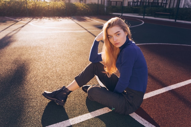 Stilvolle schöne Mädchenmodel Blondine in einem blauen Pullover sitzt auf einem Sportplatz auf dem Boden und posiert